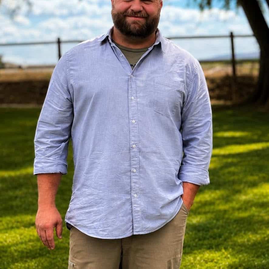 Man in casual shirt standing in a park with green grass and trees.